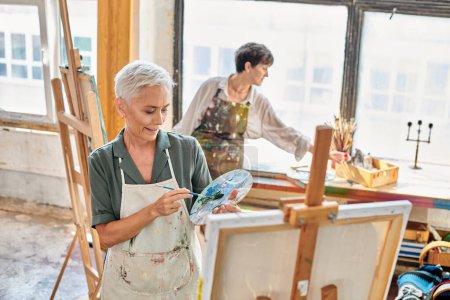 Lächeln reife Frau mischt Farben auf Farbpalette in der Nähe erfahrener Künstler in Werkstatt, Meisterkurs