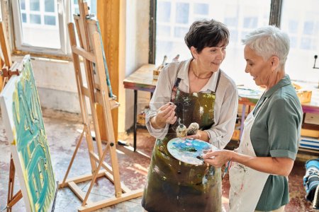 female artist showing how to mix paints on color palette to mature woman in art studio, master class