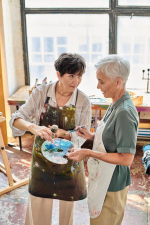 female artist mixing paints on color palette near mature woman in art studio, painting master class