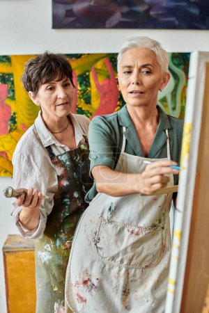 Reife Künstlerin zeigt bei Meisterkurs in moderner Handwerkswerkstatt auf Staffelei neben Frau