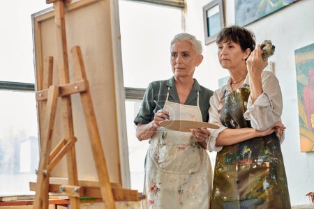 thoughtful and creative mature women looking at easel during painting master class in art workshop