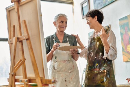 joyful mature women in aprons smiling at each other near easel during master class in art studio