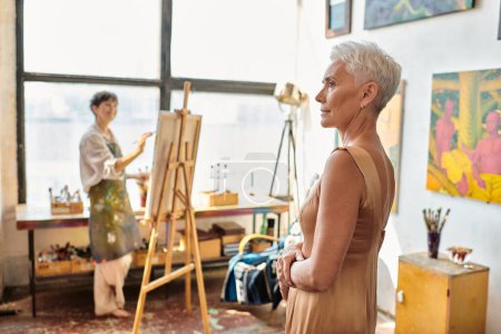 stylish mature model posing near female artist paining on easel in art workshop, creative process