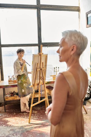 Téléchargez les photos : Gracieux modèle mature posant près artiste femme peinture sur chevalet en atelier, processus artistique - en image libre de droit