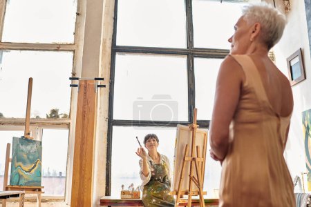 Photo for Professional mature female artist talking to model posing in art workshop, creative process - Royalty Free Image