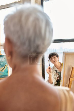 Téléchargez les photos : Artiste professionnelle de sexe féminin parlant à un modèle mature flou dans un atelier d'artisanat, processus créatif - en image libre de droit