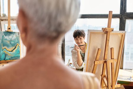 Téléchargez les photos : Joyeuse artiste féminine parlant à modèle mature floue posant dans l'atelier d'artisanat, processus créatif - en image libre de droit