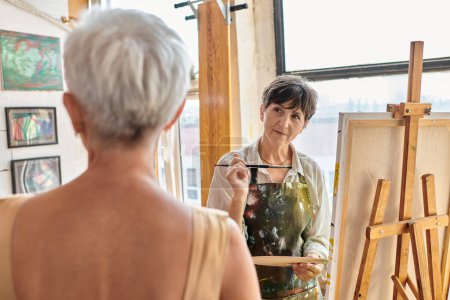 Téléchargez les photos : Artiste féminine concentrée regardant le modèle mature flou dans l'atelier d'art, processus créatif - en image libre de droit