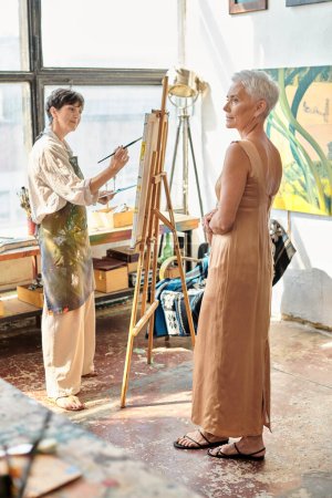talentierte Künstlerin malt reifes Modell in elegantem Kleid in Kunstwerkstatt, künstlerischer Prozess
