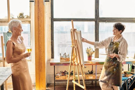 Téléchargez les photos : Heureux artiste mature peinture élégante amie posant avec verre à vin dans l'atelier d'art, créativité - en image libre de droit