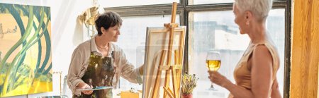 talented female artist painting elegant mature model with wine glass in art studio, banner