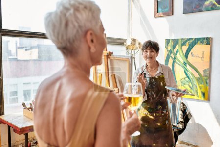 happy mature artist painting blurred female model with wine glass in art workshop, creative process