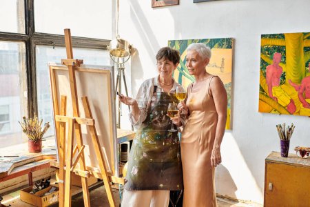 mature female artist with elegant model holding wine glasses and looking at easel in art studio