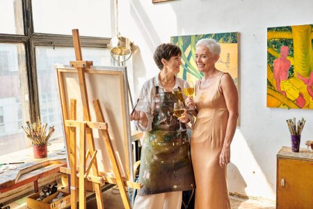 joyful female artist with elegant model standing with wine glasses near easel in art studio