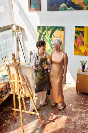 Téléchargez les photos : Artiste femme souriante avec modèle mature debout avec des verres à vin dans l'atelier d'art, vue grand angle - en image libre de droit