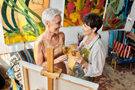 cheerful mature female friends with wine glasses near easel in art studio, artist and model teamwork