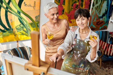 joyful mature female friends with wine glasses smiling near easel in art studio, artist and model
