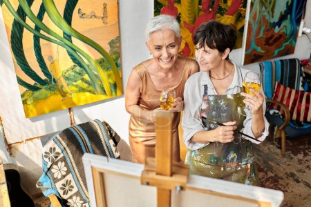 happy mature female friends with wine glasses smiling near easel in workshop, artist and model