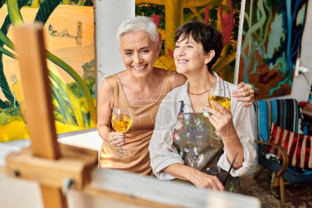 alegres mujeres maduras con copas de vino sonriendo cerca de caballete en taller de artesanía, artista y modelo
