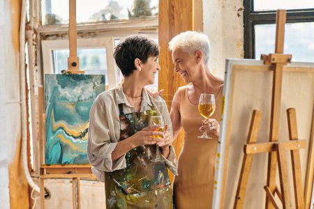 Téléchargez les photos : Joyeuse artiste féminine et élégant modèle mature avec des verres à vin riant près du chevalet en studio - en image libre de droit