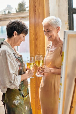 smiling female artist and elegant mature model clinking wine glasses in modern art workshop