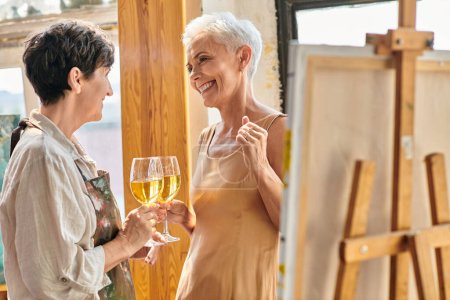 Téléchargez les photos : Heureux peintre femme et élégant modèle mature cliquetis verres à vin dans l'atelier d'art moderne - en image libre de droit