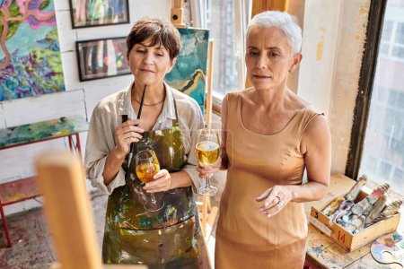 thoughtful woman artist and stylish mature model standing with wine glasses  in craft workshop