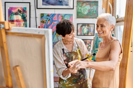 cheerful and stylish mature model with wine glass pointing at easel near woman painter in art studio