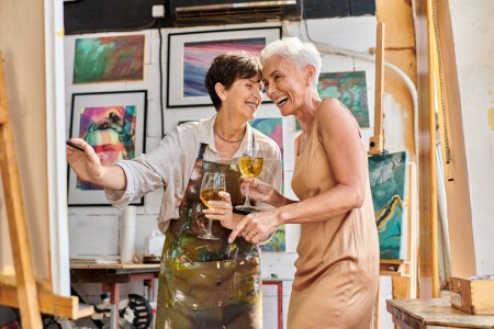 excited mature woman artist and elegant model with wine glasses laughing near easel in art studio