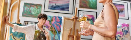 middle aged model with wine glasses posing near woman painter in art studio, horizontal banner
