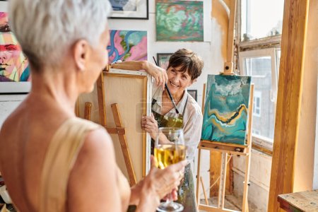 cheerful mature woman artist looking at blurred female model posing with wine glass in workshop