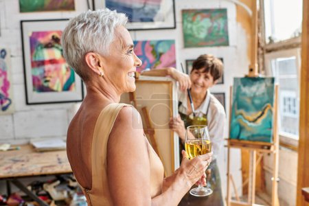 joyful and stylish woman posing with wine glass near female artist in workshop, creative process