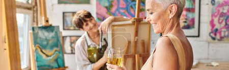stylish middle aged woman with wine glass smiling near female artist in studio, horizontal banner
