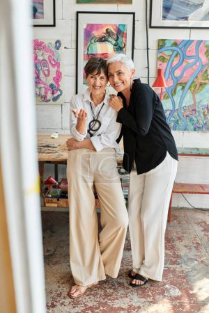 excitada artista femenina de mediana edad apuntando con la mano cerca de la pareja lesbiana en el taller de arte moderno