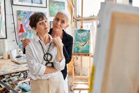 artistes lesbiennes élégantes et réfléchies regardant la peinture dans l'atelier d'artisanat moderne, l'amour et l'art