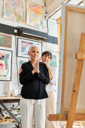 impressed female artist looking at easel near lesbian partner in workshop, teamwork and art