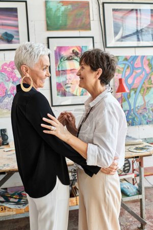 Photo for Side view of joyful and elegant lesbian couple of artists looking at each other in art workshop - Royalty Free Image