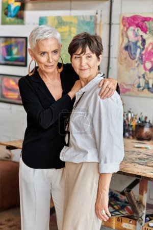 elegant and happy lesbian couple of modern artists standing and looking away in craft workshop