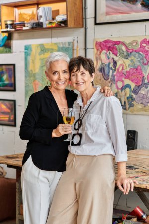 Photo for Lesbian couple of artists with wine glass embracing and looking at camera in contemporary art studio - Royalty Free Image