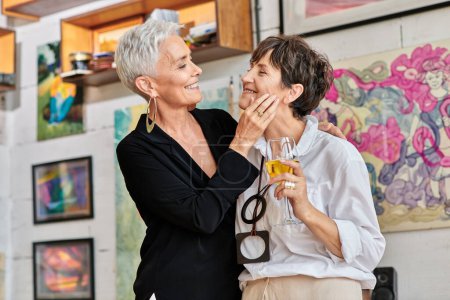 Foto de Mujer madura de moda tocando la cara de pareja lesbiana con copa de vino en el estudio de arte contemporáneo - Imagen libre de derechos