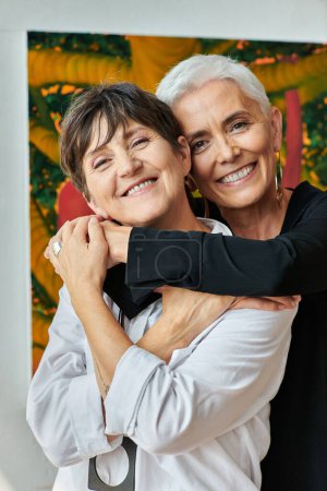 portrait d'artistes lesbiennes matures heureuses et branchées souriant à la caméra dans un atelier artisanal moderne