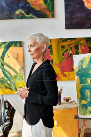 mujer artista de mediana edad de moda y confianza mirando hacia otro lado cerca de pinturas creativas en el taller