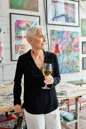 verträumte reife Künstlerin mit Weinglas im Kunstatelier mit kreativen Gemälden