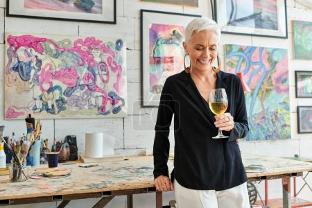 fröhliche reife Künstlerin mit Weinglas lächelnd im Kunstatelier mit kreativen Bildern
