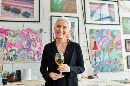 freudige reife Künstlerin mit Weinglas lächelt in Kamera im Kunstatelier mit kreativen Bildern