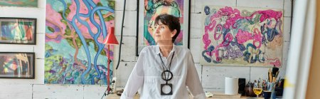 mujer madura pensativa artista de pie y mirando hacia otro lado en el estudio de arte con pinturas de colores, pancarta