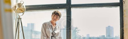 souriant mature artiste femme regardant la caméra près de la fenêtre atelier spacieux moderne, bannière