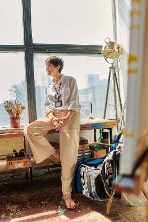 freudige reife Künstlerin sitzt am Fenster und schaut weg in moderne geräumige Werkstatt