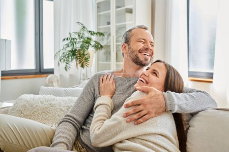excited child-free couple embracing and laughing on couch in modern living room, calm lifestyle