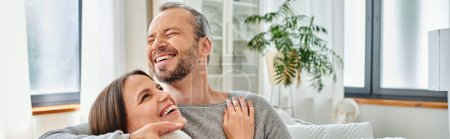 cheerful child-free couple embracing and laughing on couch in modern living room, horizontal banner